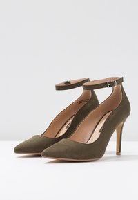 Zelené semišové lodičky s špičatou špičkou, kotníkovým páskem a jehlovým podpatkem. Interiér ve světlé barvě s minimalistickým designem.