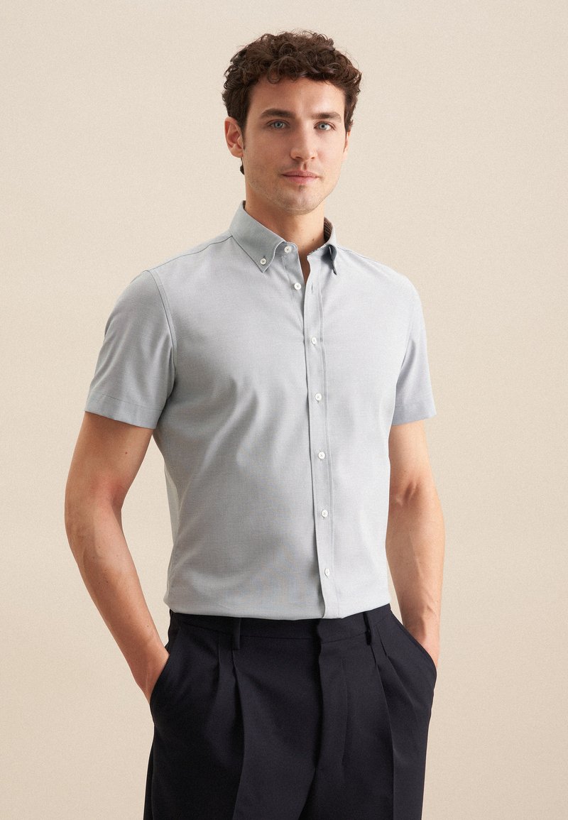 Camicia grigia chiara a maniche corte con colletto, caratterizzata da una texture liscia e una vestibilità classica, abbinata a pantaloni scuri.