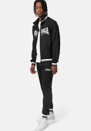 Survêtement noir avec des accents blancs, comprenant une veste zippée et des poignets élastiques, associé à un pantalon assorti et des baskets blanches.