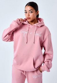 Sudadera rosa de gran tamaño hecha de tela suave, con un bolsillo canguro y cordones. En la parte frontal se muestra el texto del logo en blanco "Project x Paris."