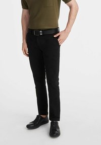 Mężczyzna stoi pewnie, ubrany w oliwkową koszulę z kr ótkim rękawem, czarne spodnie i czarne trampki, mając jedną rękę na biodrze.