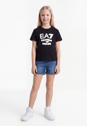 Νεαρή κοπέλα που χαμογελά, φοράει μαύρο t-shirt EA7 Emporio Armani, μπλε τζιν σορτς και λευκά ψηλά αθλητικά παπούτσια, κάθεται μπροστά σε λευκό φόντο.