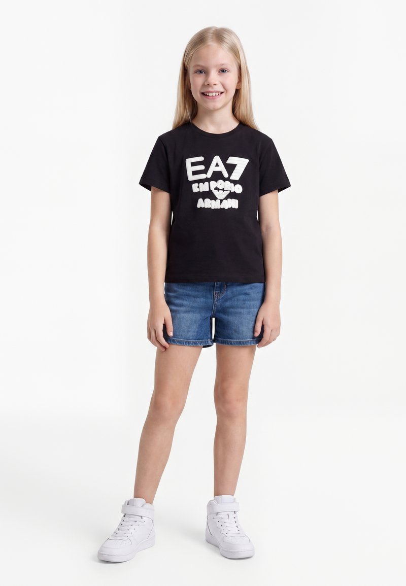 Νεαρή κοπέλα που χαμογελά, φοράει μαύρο t-shirt EA7 Emporio Armani, μπλε τζιν σορτς και λευκά ψηλά αθλητικά παπούτσια, κάθεται μπροστά σε λευκό φόντο.