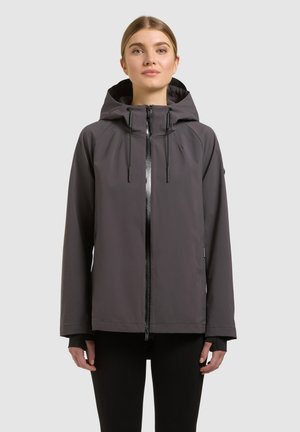 Femeie purtând o jachetă cu glugă de culoare gri închis, cu fermoar frontal și șnururi, stând pe un fundal neutru de culoare gri deschis.