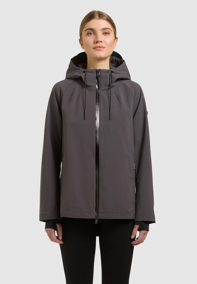 Femme portant une veste à capuche gris foncé avec une fermeture éclair frontale et des cordons, debout devant un fond gris clair neutre.