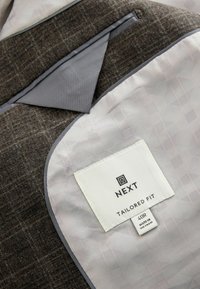 Brauner, strukturierter Stoff mit grauem Futter. Verfügt über eine dreieckige Tasche und ein Etikett mit der Aufschrift "NEXT TAILORED FIT 40R MADE IN VIETNAM".