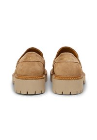 Mocassins en suédine beige avec une épaisse semelle en caoutchouc beige. Présente un détail cousu et une marque dorée sur le dessus. La vue arrière met en avant des talons arrondis.