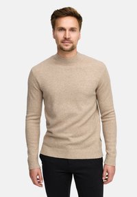 Maglione lavorato a maglia beige chiaro con scollo rotondo a costine e maniche lunghe. Caratterizzato da una texture liscia e una forma aderente. Pantaloni neri visibili.