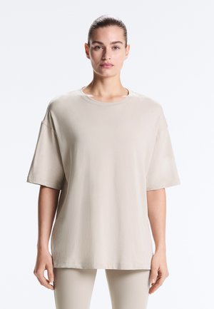 Frau mit zurückgebundenem Haar, die ein locker sitzendes beiges T-Shirt mit kurzen Ärmeln und passende hellfarbene Hosen trägt, steht vor einem weißen Hintergrund.