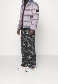 Puffa bunda v iridescentní světle fialové barvě, s předním zipem a černou logo nášivkou. Černé kalhoty s vzorem obsahují opakující se grafické designy. Bílé tenisky.