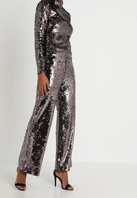 Pailletten-Outfit in metallischen Schattierungen von Silber und Schwarz. Mit langen Ärmeln, einem hohen Ausschnitt und weit geschnittenen Hosen. Schwarze Sandalen mit Absatz.