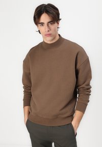 Brown sweatshirt with a high neckline and ribbed cuffs, featuring a subtle logo detail at the hem. Material appears soft and textured.