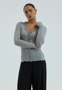 Femme aux cheveux foncés portant un haut gris ajusté à manches longues avec des bords à volants et un pantalon noir ample, debout devant un fond uni.
