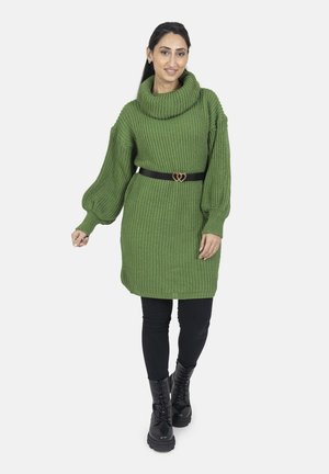 Vrouw draagt een groene gebreide sweaterjurk, zwarte riem met hartvormige gesp, zwarte legging en zwarte veterlaarzen, staand op een witte achtergrond.