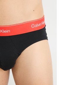 Fekete pamut alsónadrág piros elasztikus derékrésszel, amelyen a "Calvin Klein" logó fehér színben látható. Sima textúra és illeszkedő kialakítás.
