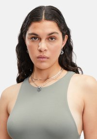 Silberne Kettenkette mit einem herzförmigen Anhänger, kombiniert mit passenden quadratischen Ohrringen. Modell trägt ein enges olivgrünes Oberteil.