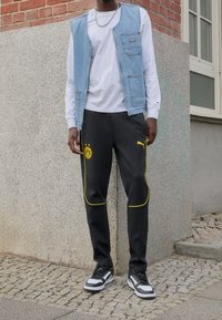 Man in zwarte en gele Puma Borussia Dortmund sportbroek, witte lange mouwen shirt, lichtblauwe spijkerjasje, en zwart-witte sneakers.