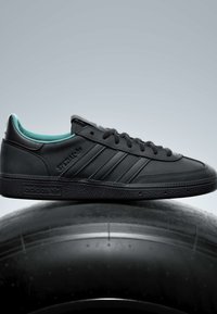 Baskets noires Adidas Spezial à trois bandes et design à lacets, posées sur un grand pneu noir sur fond gris uni.