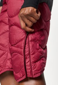 Χέρι που κρατάει το φερμουάρ σε μπορντό επενδυμένη φούστα, με μέρος του μηρού και μαύρου μανικιού ορατού σε λευκό φόντο.