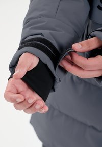 Grå puffjakkeærme med et sort elastisk indre lag og velcro-rem. Hænder justerer ærmet, hvilket afslører en glat struktur og fleksibel pasform.