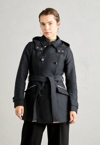 Zwarte waterdichte trenchcoat met een dubbelzijdig ontwerp, capuchon, riem en ritszakken aan de zijkant. Met metalen accenten. Gladde textuur.