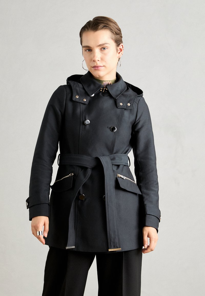 Zwarte waterdichte trenchcoat met een dubbelzijdig ontwerp, capuchon, riem en ritszakken aan de zijkant. Met metalen accenten. Gladde textuur.