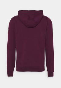 Huvtröja i mörklila tyg med långa ärmar och ribbade muddar. Inkluderar en logotyp på vänster axel.