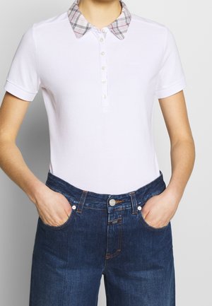 Personne portant un polo blanc à manches courtes avec un col à carreaux et un jean en denim bleu, les mains dans les poches, debout devant un fond uni.