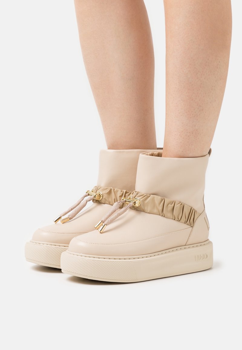Bottes beiges à cheville avec semelles épaisses et bandes en tissu froncé maintenues par des boutons dorés, portées sur des jambes nues sur fond blanc.
