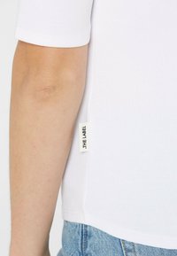 Weißes geripptes T-Shirt mit kurzen Ärmeln, das an der Seite ein kleines Stoffetikett hat. Das Shirt hat eine lockere Passform und eine glatte Textur.