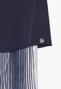 Námořnická modrá blůza s hladkou texturou, zdobená stříbrným akcentem v podobě knoflíku, přesažená přes látku s bílými a námořnickými pruhy.