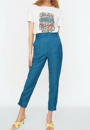 Vrouw draagt een wit T-shirt met print, hoge blauwe broek met knopen en gele open teen sandalen met hak, staand tegen een witte achtergrond.