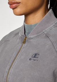 Giacca bomber grigia con chiusura a zip, collo a costine e dettaglio del logo ricamato. Caratterizzata da una texture morbida e un design casual.