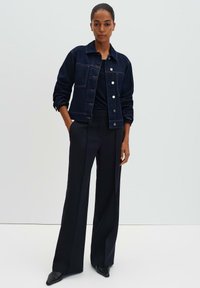 Giacca in denim scuro con bottoni a pressione, tasche laterali e maniche arrotolate; abbinata a pantaloni neri a vita alta e scarpe nere appuntite.