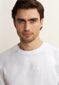 Maglietta bianca in cotone con scollatura rotonda, caratterizzata da un sottile testo in rilievo sul petto e una superficie liscia.