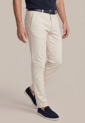 Man draagt beige broek, zwarte riem, marineblauwe loafers en een donker marineblauw overhemd, staand tegen een effen beige achtergrond.