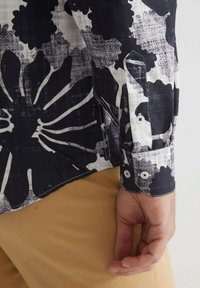 Chemise à manches longues avec un motif floral noir et blanc, poignets boutonnés et tissu texturé. Portée avec un pantalon de couleur claire.