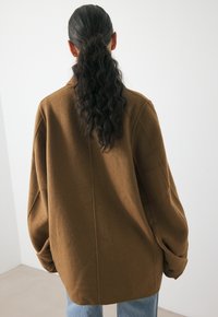 Brown wool coat with an oversized fit, featuring a notched collar and large, slouchy sleeves. Textured fabric and simple button detail.