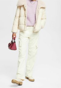 Veste matelassée beige clair avec col en fausse fourrure, pull en maille violet pastel, pantalon en velours côtelé crème, et petit sac à main bordeaux. Sabots beige.