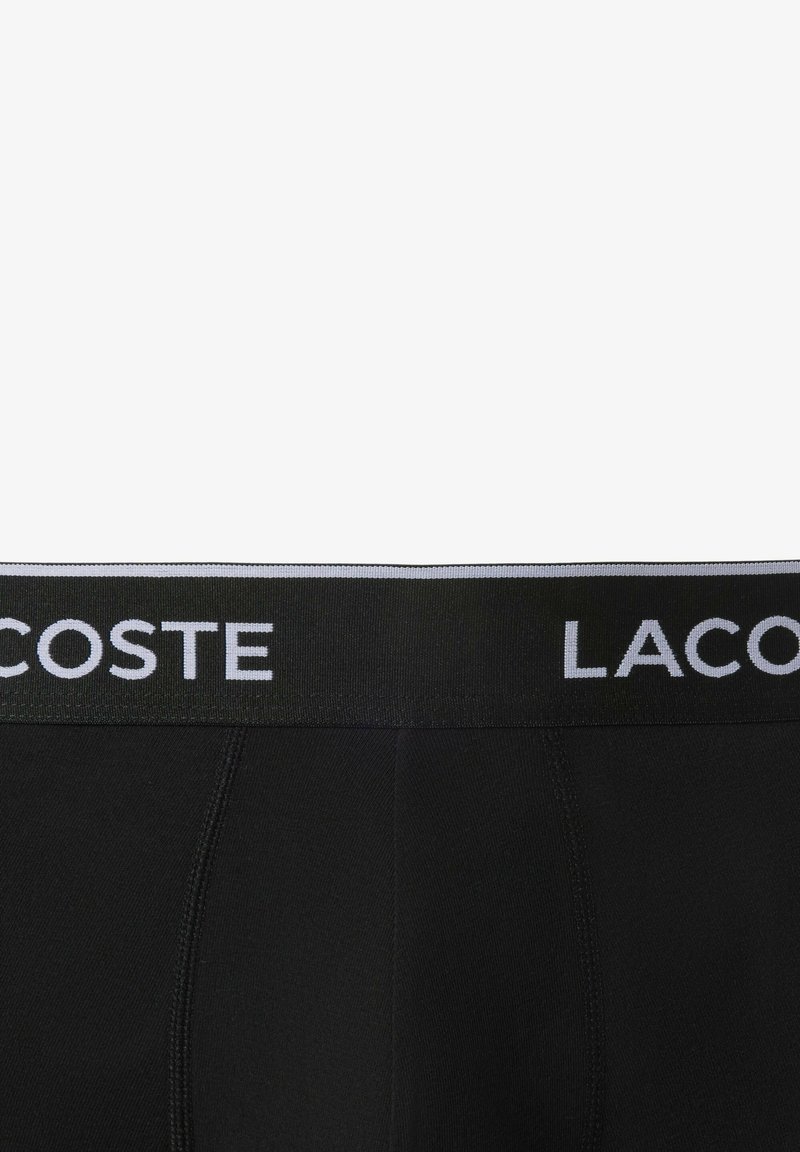 Schwarze Boxerbriefs mit einem weichen elastischen Bund, der den weißen Text "LACOSTE" trägt. Glatter Stoff mit einer körpernahen Passform.