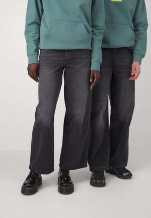 Jeans anchos negros con un acabado desgastado, hechos de mezclilla. Combinados con botas oscuras y una sudadera de color verde azulado con un bolsillo delantero.