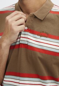 Polo marron à rayures horizontales rouges et bleues. Comprend un col avec boutons et un logo brodé "Original" au-dessus de la zone de la poche.