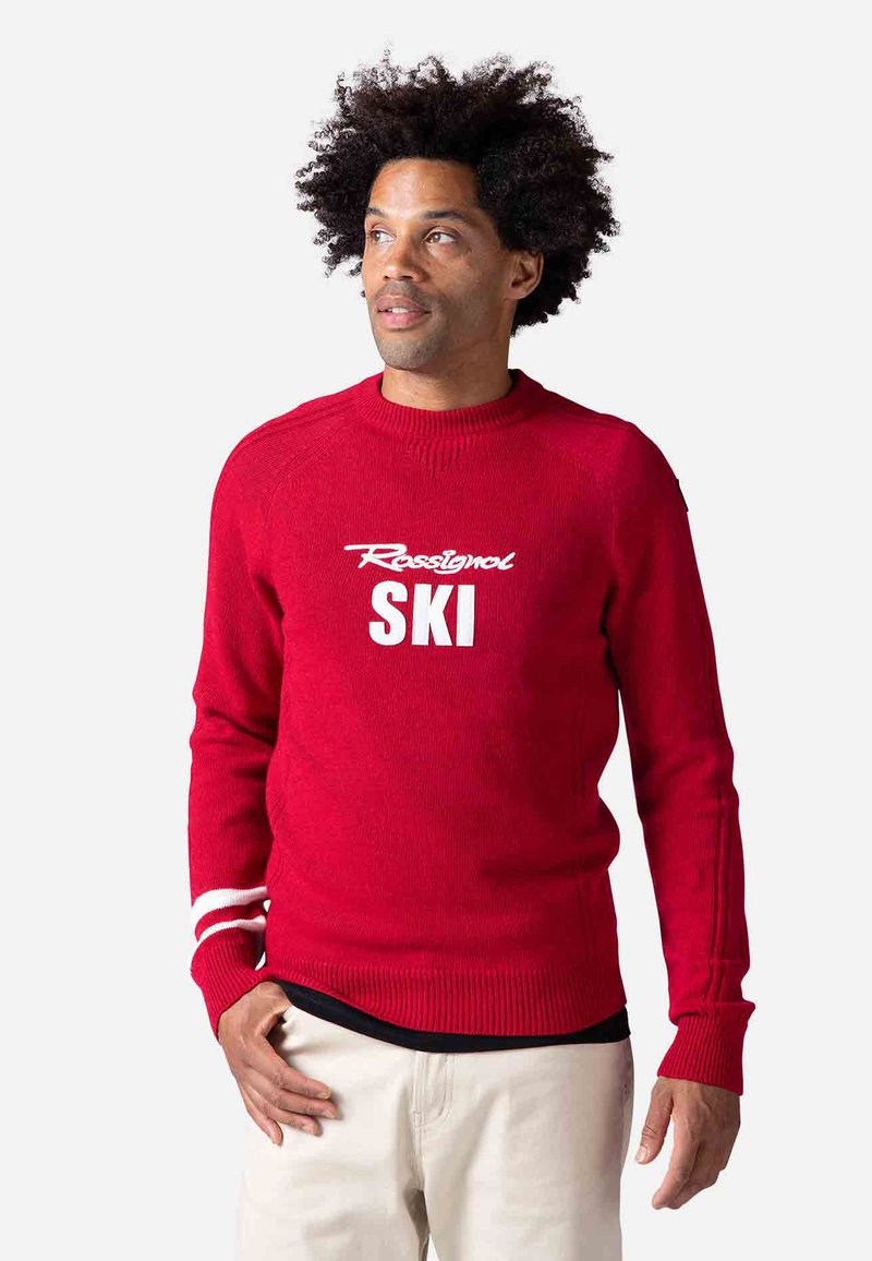 Roter Strickpullover mit Rundhalsausschnitt, weißem "Rassignol" und "SKI" Druck sowie weißen Streifen an den Ärmeln. Rippbündchen und Saum.