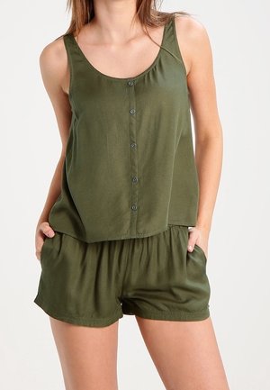 Frau trägt ein olivgrünes ärmelloses, geknöpftes Oberteil und passende weite Shorts, steht mit den Händen in den Taschen vor einem schlichten Hintergrund.