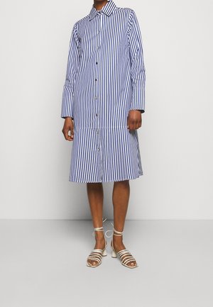 Abito camicia a righe blu e bianche, con bottone frontale, maniche lunghe e gonna svasata. Abbinato a sandali bianchi con cinturini.