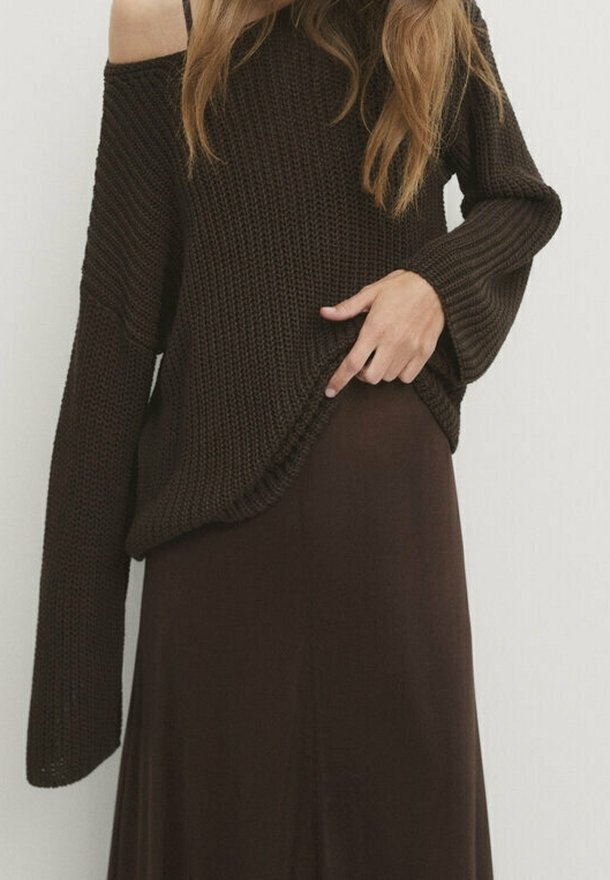 Femme portant un pull en grosse maille marron foncé à épaules dénudées avec des manches longues et une jupe assortie marron foncé, tirant le bas du pull à la main.