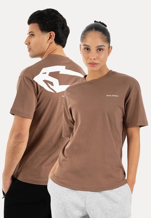 Mężczyzna i kobieta w brązowych koszulkach, kobieta zwrócona przodem z rękami w kieszeniach, mężczyzna obrócony bokiem, pokazujący biały nadruk na plecach.