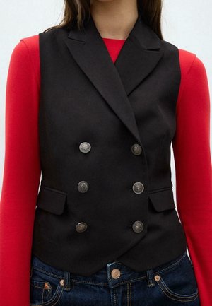 Femme portant un gilet noir sans manches à double boutonnage avec des boutons argentés sur un haut rouge à manches longues et un jean bleu.