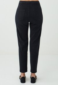 Pantalons noirs taille haute portés par une personne, vus de dos. L'ensemble est associé à des sandales à lanières noires. Fond blanc uni.
