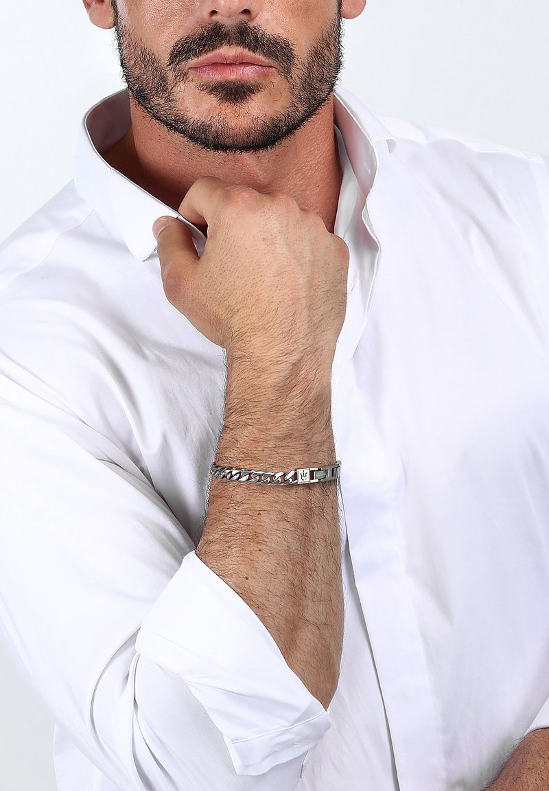 Silverfärgad kedjearmband med rektangulära länkar och en låsning, bärs på ett handled. Modellen har på sig en vit kragblus, delvis uppvikt.